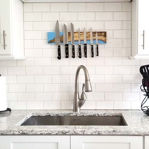 Magnetic knife rack holding kitchen knives above a sink in a white and gray kitchen. Handcrafted with spalted live edge maple wood and carolina blue epoxy resin.